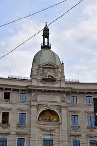 A historic European building with ornate architectural details, including a prominent dome with a decorative spire and windows. The facade features intricate stone carvings and a colorful fresco or mosaic beneath the central arch. Several windows are symmetrically placed, and the words 'ASSICURAZIONI GENERALI' are written above the fresco. Overhead wires crisscross the sky above the building.