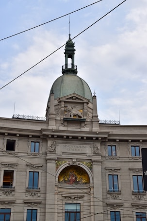 A historic European building with ornate architectural details, including a prominent dome with a decorative spire and windows. The facade features intricate stone carvings and a colorful fresco or mosaic beneath the central arch. Several windows are symmetrically placed, and the words 'ASSICURAZIONI GENERALI' are written above the fresco. Overhead wires crisscross the sky above the building.