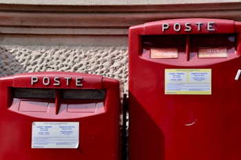 Two red Italian post boxes with text reading 'POSTE' at the top and informational stickers detailing collection times. The background is a textured stone wall.