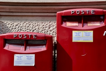 Two red Italian post boxes with text reading 'POSTE' at the top and informational stickers detailing collection times. The background is a textured stone wall.