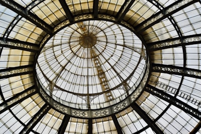 A circular glass ceiling with an intricate wrought iron framework creating a pattern of concentric rings. The structure includes detailed metalwork and is composed of transparent and opaque glass panels.