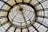 A circular glass ceiling with an intricate wrought iron framework creating a pattern of concentric rings. The structure includes detailed metalwork and is composed of transparent and opaque glass panels.
