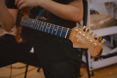 A person is holding an electric guitar, focusing on the guitar's neck and tuning pegs. The environment seems to be indoors, possibly in a music studio or practice room.