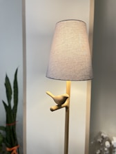 A wooden floor lamp with a neutral-colored lampshade. The lamp features a decorative wooden bird perched on the stand, creating a minimalist and natural aesthetic. In the background, there is a snake plant with several tall green leaves.