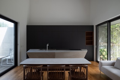 A modern kitchen space features a minimalist design with a large island counter. The cabinetry is dark and sleek, complemented by a stylish faucet. Wooden flooring harmonizes with the wooden dining set, consisting of a table and several chairs. Wide glass doors on either side offer ample natural light and views of the outdoors, with a patio visible on one side.
