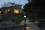A modern house with illuminated exterior lights during dusk. The house is surrounded by a stone wall and lush greenery. A camera on a tripod is positioned in the foreground, aimed at the house. The sky is slightly overcast, adding a serene ambiance. Some buildings are visible in the background, and the pathway leading to the house is nicely lit with ground lights.