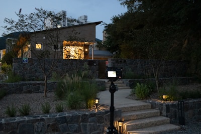 A modern house with illuminated exterior lights during dusk. The house is surrounded by a stone wall and lush greenery. A camera on a tripod is positioned in the foreground, aimed at the house. The sky is slightly overcast, adding a serene ambiance. Some buildings are visible in the background, and the pathway leading to the house is nicely lit with ground lights.