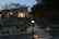 A modern house with illuminated exterior lights during dusk. The house is surrounded by a stone wall and lush greenery. A camera on a tripod is positioned in the foreground, aimed at the house. The sky is slightly overcast, adding a serene ambiance. Some buildings are visible in the background, and the pathway leading to the house is nicely lit with ground lights.