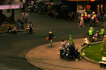 A bustling street scene featuring multiple people, some wearing traditional hats, and riding motorcycles. There are street vendors with fruit stalls and a variety of goods visible on the sidewalks. The area is lively with people walking, riding bikes, and engaging in daily activities, showcasing a typical market atmosphere.