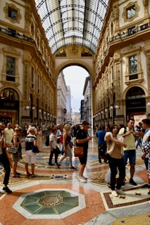 A bustling indoor shopping arcade with an ornate glass ceiling, detailed architectural features, and a mosaic floor. People are walking, shopping, and engaging in conversations. High-end stores like Dior and Prada line the sides.
