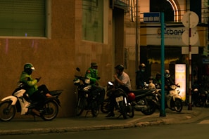 A group of motorcyclists, some wearing helmets and reflective jackets, are gathered on the sidewalk near a building at night. The setting is urban with a sign indicating a street name. One person is interacting with a phone, while others appear to be in a casual conversation. The area is dimly lit with artificial street lighting giving a warm tone to the scene.