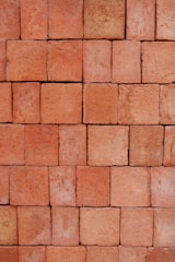 A neatly stacked arrangement of uniform red bricks arranged in a tight pattern covering the entire frame. The bricks vary slightly in hue and texture, giving the arrangement a somewhat rustic and natural appearance.