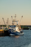 A fishing boat docked at a harbor with other boats visible in the background. The boat is painted blue and white with the code SC 12 on its bow. The surrounding environment includes calm water and a shoreline with grassy areas and some buildings in the distance.