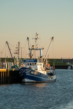 A fishing boat docked at a harbor with other boats visible in the background. The boat is painted blue and white with the code SC 12 on its bow. The surrounding environment includes calm water and a shoreline with grassy areas and some buildings in the distance.