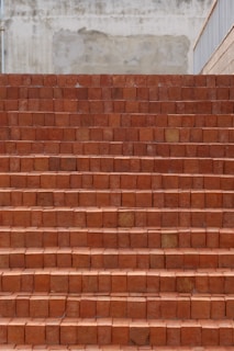 A set of brick stairs constructed from reddish-brown bricks, leading upward. The surface behind the stairs appears to be a textured and slightly weathered wall in shades of beige. The right side features a railing.