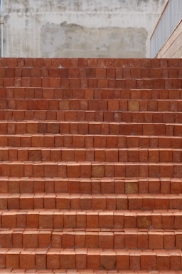 A set of brick stairs constructed from reddish-brown bricks, leading upward. The surface behind the stairs appears to be a textured and slightly weathered wall in shades of beige. The right side features a railing.
