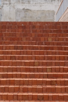 A set of brick stairs constructed from reddish-brown bricks, leading upward. The surface behind the stairs appears to be a textured and slightly weathered wall in shades of beige. The right side features a railing.