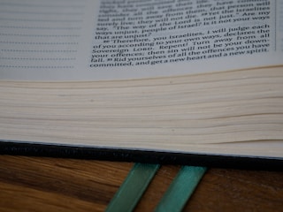 A close-up of an open book with visible text and neatly aligned pages. Two green ribbon bookmarks extend from the bottom, resting on a wooden surface.