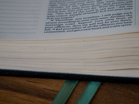 A close-up of an open book with visible text and neatly aligned pages. Two green ribbon bookmarks extend from the bottom, resting on a wooden surface.