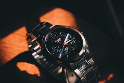 A metallic wristwatch with a prominent bezel and a black and blue dial. The watch face displays analog time with three sub-dials, indicating additional functions like chronograph. Stainless steel links form the bracelet, reflecting ambient light and creating highlights and shadows.
