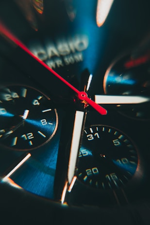 Close-up view of an intricate watch face featuring multiple dials and a vibrant red second hand. The watch has a metallic finish with visible branding.