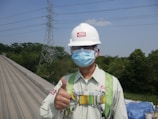 A person wearing safety equipment including a helmet and mask gives a thumbs-up on a rooftop. In the background, there are tall trees and a high-voltage power line tower against a clear blue sky.