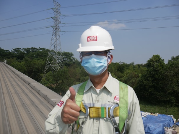 A person wearing safety equipment including a helmet and mask gives a thumbs-up on a rooftop. In the background, there are tall trees and a high-voltage power line tower against a clear blue sky.