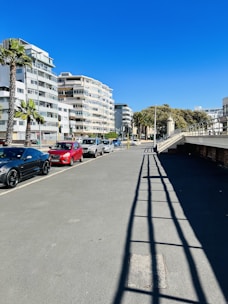 A street scene with modern apartment buildings on the left and several parked cars, including a black sports car and a red vehicle. Palm trees are lining the sidewalk, and the sky is clear and blue. The shadows of railings create patterns on the pavement.