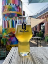 A glass of beer with a logo, placed on a wooden table in an outdoor setting. In the background, a large structure is painted with a vivid geometric and colorful pattern. The atmosphere is bright and possibly part of an open-air establishment with additional tables and chairs.