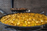 A large pan filled with traditional paella, featuring vibrant yellow rice, chicken pieces, and assorted vegetables. Another pan in the background contains seafood paella with mussels and prawns. The pans are set against a brick wall.