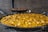 A large pan filled with traditional paella, featuring vibrant yellow rice, chicken pieces, and assorted vegetables. Another pan in the background contains seafood paella with mussels and prawns. The pans are set against a brick wall.
