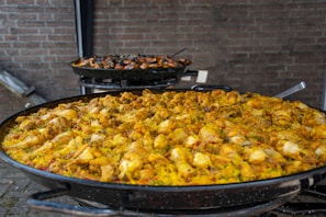 A large pan filled with traditional paella, featuring vibrant yellow rice, chicken pieces, and assorted vegetables. Another pan in the background contains seafood paella with mussels and prawns. The pans are set against a brick wall.