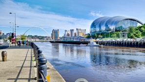 A modern architectural scene featuring a river with a bridge crossing over it. On the right, a large, curved glass building sits on a grassy incline, and several tall, colorful buildings are visible in the background. People are walking and cycling along a paved riverside path lined with lampposts.
