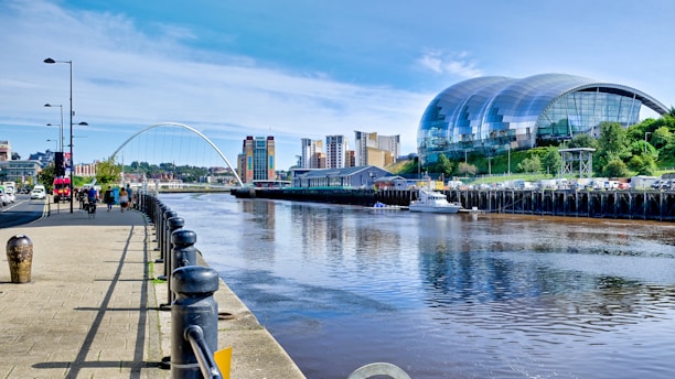 A modern architectural scene featuring a river with a bridge crossing over it. On the right, a large, curved glass building sits on a grassy incline, and several tall, colorful buildings are visible in the background. People are walking and cycling along a paved riverside path lined with lampposts.