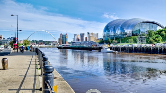 A modern architectural scene featuring a river with a bridge crossing over it. On the right, a large, curved glass building sits on a grassy incline, and several tall, colorful buildings are visible in the background. People are walking and cycling along a paved riverside path lined with lampposts.