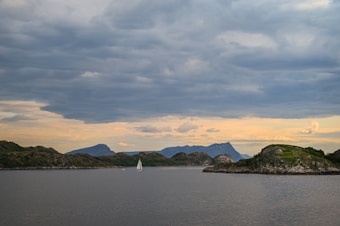 A serene seascape featuring a sailboat gliding on calm waters under a sky filled with dramatic, layered clouds. The horizon showcases rugged, green-topped hills and distant mountains, creating a tranquil yet majestic atmosphere.