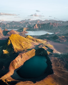 A breathtaking aerial view of a volcanic landscape with a large crater filled with deep blue water. The surrounding terrain features rugged hills and mountains, with varying shades of green, brown, and orange, suggesting different types of vegetation and rock. The sky is clear with soft clouds, casting gentle shadows over the landscape.