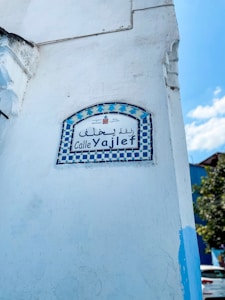 A white wall featuring a decorative ceramic street sign with a checkered blue and white border. The sign displays text in both Arabic and Latin script, reading 'Calle Yajlef'. A blue sky and some greenery are visible in the background.