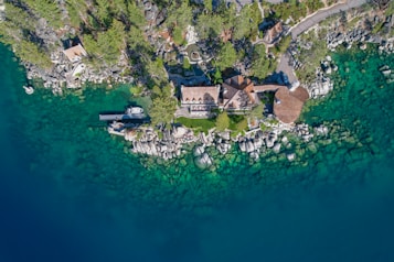 Aerial view of a lakeside property surrounded by lush greenery and rocky terrain. A boathouse and dock are visible, with crystal-clear turquoise water along the shoreline. The house is nestled among trees and connected to a road leading in and out of the area.