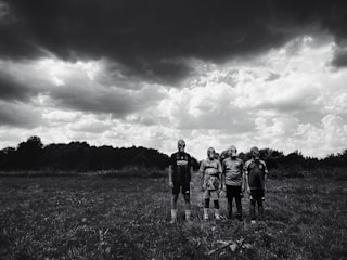 Four people stand in an open grassy field wearing gas masks under a dramatic, cloudy sky. The dark clouds create a stark contrast with the lighter areas of the sky, enhancing the mysterious and eerie atmosphere.