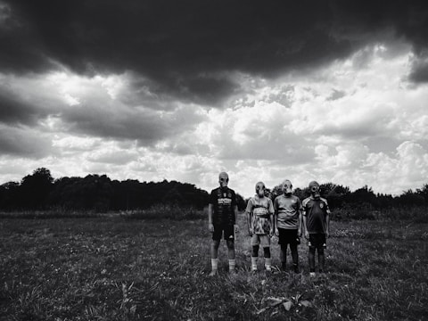 Four people stand in an open grassy field wearing gas masks under a dramatic, cloudy sky. The dark clouds create a stark contrast with the lighter areas of the sky, enhancing the mysterious and eerie atmosphere.