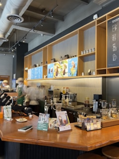 A cozy café interior with a wooden counter displaying various coffee-making equipment and promotional materials. Baristas in uniforms work behind the counter, and the wall features shelving with coffee-related items and a menu board above.