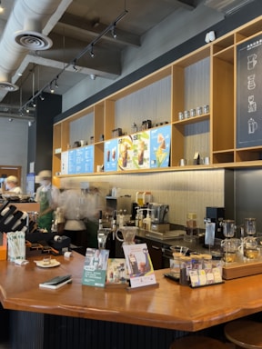 A cozy café interior with a wooden counter displaying various coffee-making equipment and promotional materials. Baristas in uniforms work behind the counter, and the wall features shelving with coffee-related items and a menu board above.