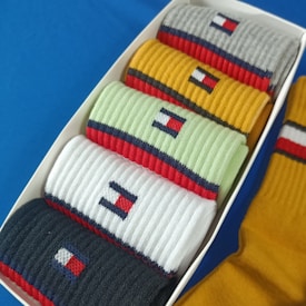 A set of folded socks in different colors arranged neatly in a box. Each sock features a detailed pattern with vibrant stripes, including elements of red, white, and blue, against background colors of grey, yellow, mint, white, and black.
