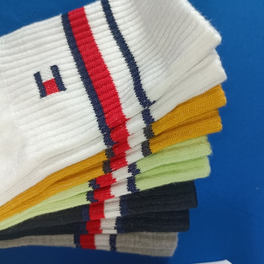 A collection of colorful socks neatly stacked on top of a blue surface. The socks display a variety of colors including white, yellow, green, black, and gray, each featuring a red and navy blue striped pattern. One sock has a small embroidered logo.