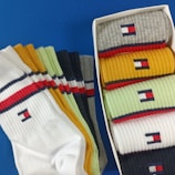 A collection of colorful socks with a distinctive logo is displayed. The socks are arranged both inside a box and spread out beside it. Various colors like white, yellow, green, gray, and blue can be seen, each featuring a small embroidered logo near the top. The background is a solid blue surface.