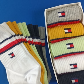 A collection of colorful socks with a distinctive logo is displayed. The socks are arranged both inside a box and spread out beside it. Various colors like white, yellow, green, gray, and blue can be seen, each featuring a small embroidered logo near the top. The background is a solid blue surface.