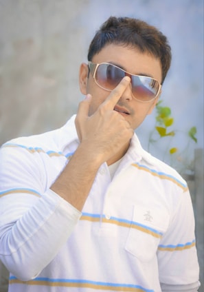 A man is posing with sunglasses, wearing a white polo shirt with blue and orange stripes. His finger is touching the bridge of the sunglasses, and there's a blurred plant in the background.