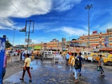 A bustling city scene with people walking on a wet street, surrounded by vibrant yellow taxis and vehicles. Buildings in the background display various advertisements and signs. The sky is a bright blue with scattered clouds, and a large Indian flag is mounted on a tall pole.