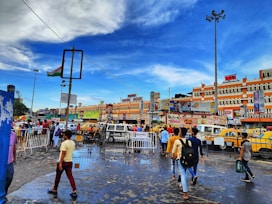A bustling city scene with people walking on a wet street, surrounded by vibrant yellow taxis and vehicles. Buildings in the background display various advertisements and signs. The sky is a bright blue with scattered clouds, and a large Indian flag is mounted on a tall pole.